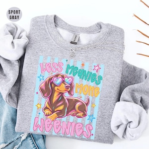 May include: A sport gray crewneck sweatshirt featuring a graphic of a brown dachshund wearing pink heart-shaped sunglasses. The text reads Less Meanies More Weenies in pink and teal lettering, surrounded by small colorful stars on a striped background.