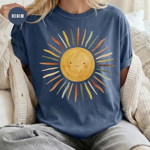 May include: A denim blue t-shirt featuring a central graphic of a yellow sun with a simple smiling face. The sun is surrounded by radiating rays in various muted tones of orange, green, and brown, creating a colorful and distinct circular pattern.