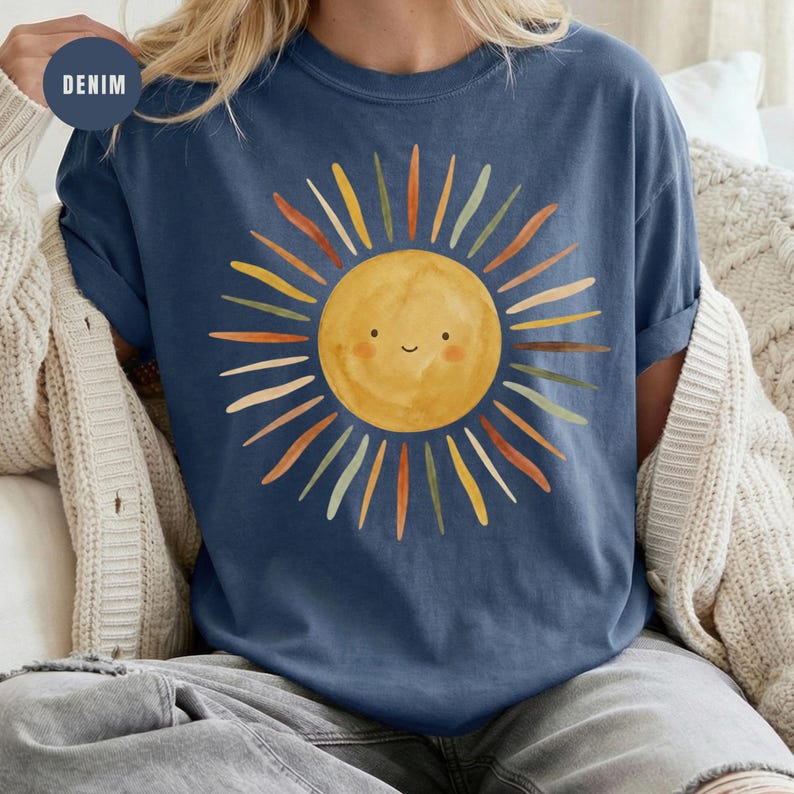 May include: A denim blue t-shirt featuring a central graphic of a yellow sun with a simple smiling face. The sun is surrounded by radiating rays in various muted tones of orange, green, and brown, creating a colorful and distinct circular pattern.
