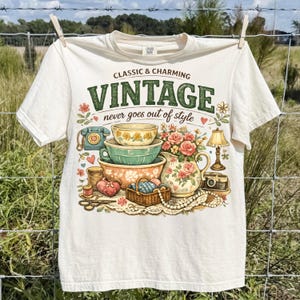 May include: A cream-colored t-shirt featuring a graphic print with the text "CLASSIC & CHARMING VINTAGE never goes out of style." The illustration includes stacked mixing bowls, a floral pitcher, a rotary phone, a lamp, and sewing supplies.