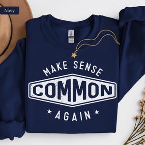 May include: A navy blue crewneck sweatshirt featuring a white graphic design with the text "MAKE SENSE COMMON AGAIN" enclosed in a diamond-shaped border. The design includes two small stars and a distressed texture. A gold star pendant necklace rests on the collar.