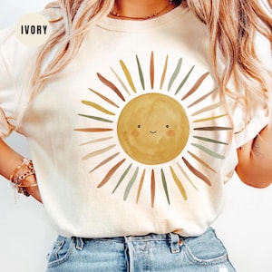 May include: An ivory crewneck t-shirt featuring a central graphic of a smiling sun with a watercolor texture. The sun is surrounded by radiating rays in muted shades of yellow, brown, and sage green. The word IVORY is printed in the top left corner.