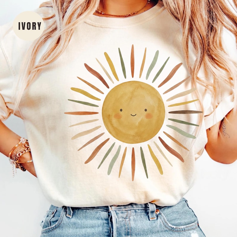 May include: An ivory crewneck t-shirt featuring a central graphic of a smiling sun with a watercolor texture. The sun is surrounded by radiating rays in muted shades of yellow, brown, and sage green. The word IVORY is printed in the top left corner.
