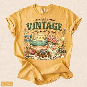 May include: A mustard yellow short-sleeve t-shirt featuring a graphic print with the text CLASSIC & CHARMING VINTAGE never goes out of style. The illustration includes stacked bowls, a floral pitcher, a rotary phone, a camera, and sewing items.
