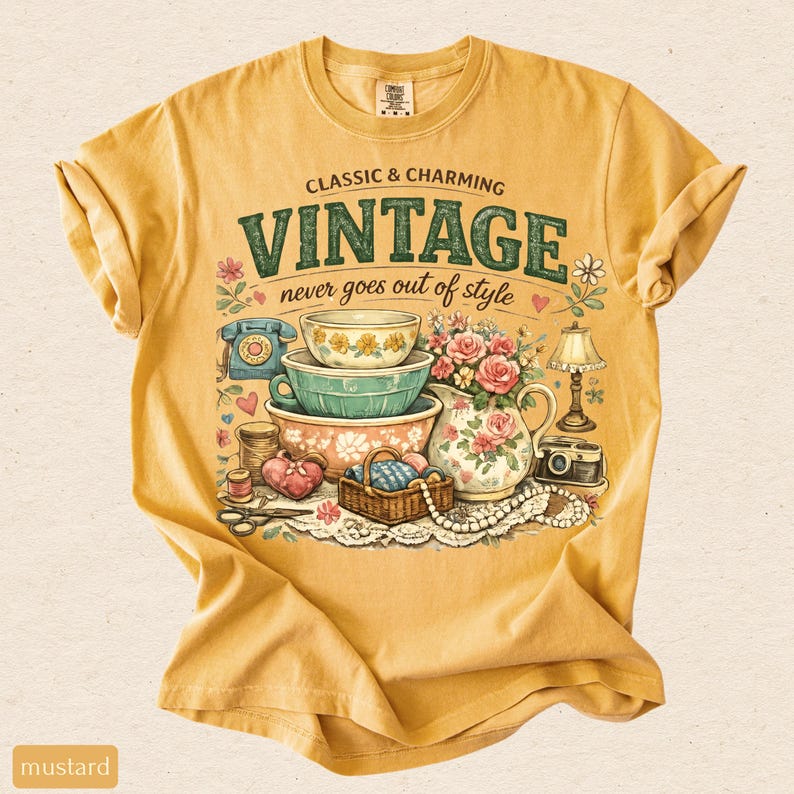 May include: A mustard yellow short-sleeve t-shirt featuring a graphic print with the text CLASSIC & CHARMING VINTAGE never goes out of style. The illustration includes stacked bowls, a floral pitcher, a rotary phone, a camera, and sewing items.