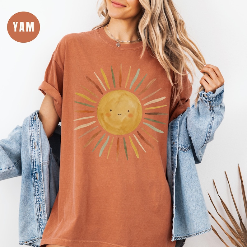 May include: A rust-colored short-sleeve t-shirt featuring a central graphic of a smiling yellow sun with radiating rays in shades of brown, green, and yellow. The shirt is layered under a light blue denim jacket, creating a casual, textured appearance.
