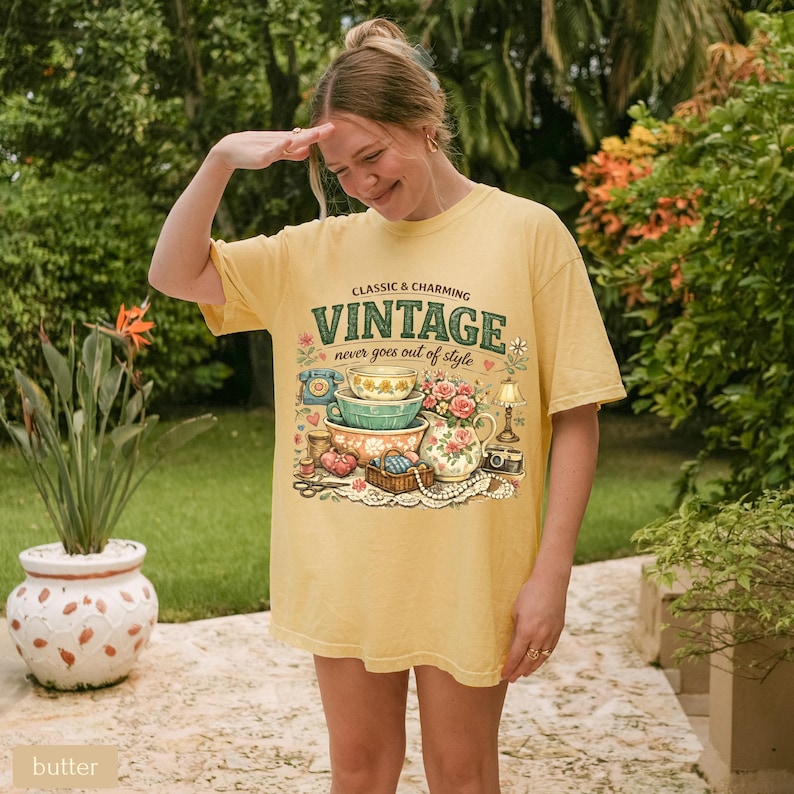 May include: A butter yellow t-shirt featuring a graphic print with the text CLASSIC & CHARMING VINTAGE never goes out of style. The illustration includes stacked bowls, a floral pitcher, a rotary phone, a lamp, and sewing supplies.