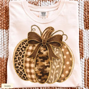 May include: An ivory t-shirt featuring a large pumpkin graphic with a brown bow. The pumpkin segments display various patterns including leopard print, brown plaid, camouflage, a leafy vine design, and white polka dots on a tan background.