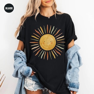 May include: A black t-shirt featuring a central graphic of a yellow sun with a simple smiling face. The sun is surrounded by radiating rays in various muted tones including orange, green, and brown. The shirt has short sleeves and a crew neckline.
