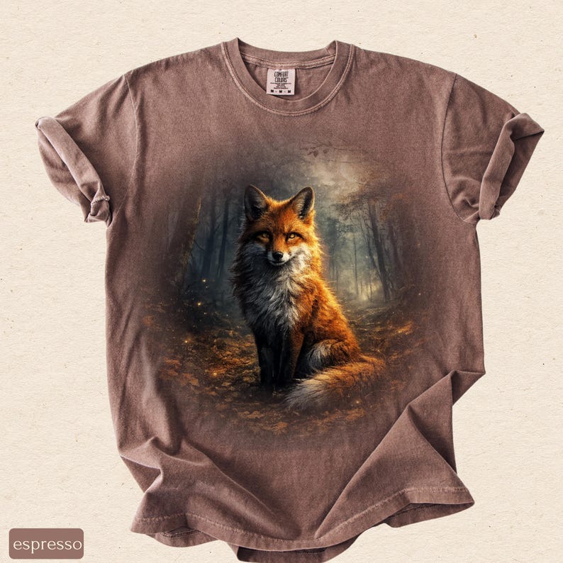 May include: Espresso-coloured t-shirt with a detailed illustration of a fox in a forest. The fox has orange and white fur, set against a backdrop of trees and foliage. The shirt has a crew neck and short sleeves.
