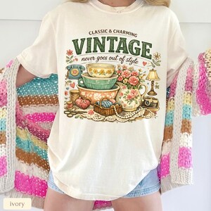 May include: An ivory t-shirt featuring a graphic print with the text "CLASSIC & CHARMING VINTAGE never goes out of style". The illustration includes stacked bowls, a floral pitcher, a rotary phone, a lamp, a camera, and a basket of yarn.