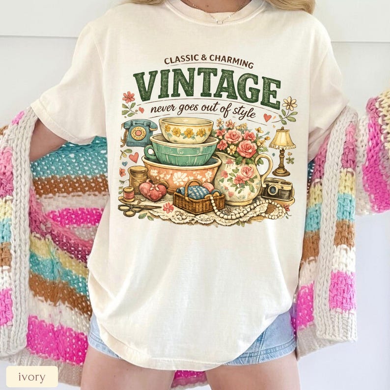 May include: An ivory t-shirt featuring a graphic print with the text "CLASSIC & CHARMING VINTAGE never goes out of style". The illustration includes stacked bowls, a floral pitcher, a rotary phone, a lamp, a camera, and a basket of yarn.