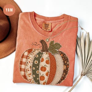 May include: A burnt orange short-sleeve t-shirt featuring a large, central pumpkin graphic composed of various fabric patches in plaid, floral, and solid patterns. The shirt has a crew neckline and a Comfort Colors brand label at the inner collar.
