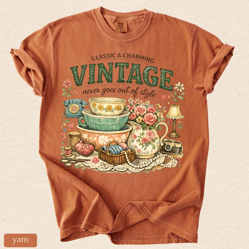 May include: A rust-colored t-shirt featuring a graphic print with the text "CLASSIC & CHARMING VINTAGE never goes out of style". The illustration includes stacked bowls, a floral pitcher, a rotary phone, a lamp, a camera, and a sewing basket.