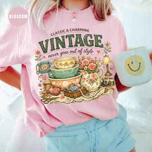 May include: A light pink t-shirt featuring a graphic print with the text "CLASSIC & CHARMING VINTAGE never goes out of style". The illustration includes stacked bowls, a floral pitcher, a telephone, a lamp, a basket, and a string of pearls.