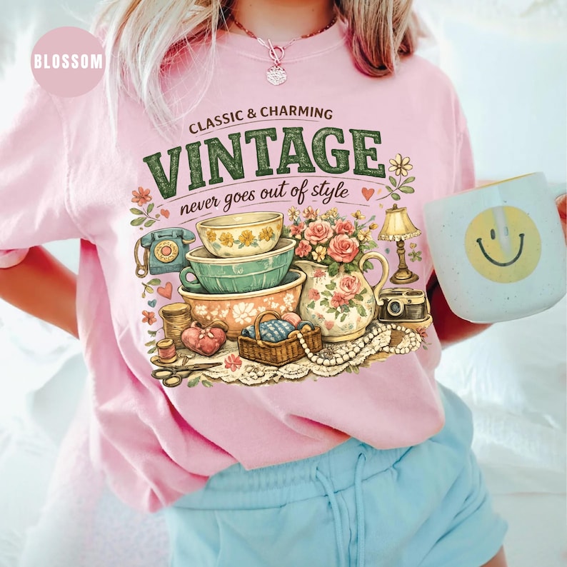 May include: A light pink t-shirt featuring a graphic print with the text "CLASSIC & CHARMING VINTAGE never goes out of style". The illustration includes stacked bowls, a floral pitcher, a telephone, a lamp, a basket, and a string of pearls.