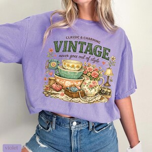 May include: A violet t-shirt featuring a graphic print with the text "CLASSIC & CHARMING VINTAGE never goes out of style". The illustration includes stacked bowls, a floral pitcher, a rotary phone, a camera, a lamp, and a sewing basket.