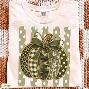 May include: An ivory t-shirt featuring a graphic of a pumpkin with a green bow. The pumpkin segments display various patterns including leopard print, buffalo check, camouflage, and white polka dots. The graphic is set against vertical olive stripes.