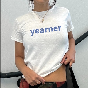 May include: A white short-sleeved t-shirt featuring the word "yearner" printed in a light blue sans-serif font across the chest. The garment has a classic crew neckline and is worn with two layered gold-toned necklaces and gold-toned rings.