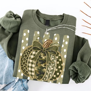 May include: An olive green crewneck sweatshirt featuring a central graphic of a pumpkin with varied patterns including leopard print, plaid, camouflage, and polka dots. The pumpkin is topped with a large olive green bow and a gold stem.
