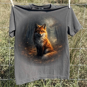 May include: A dark grey t-shirt displaying a detailed illustration of a fox with orange and white fur, set against a forest backdrop. The fox is sitting on a bed of leaves. The t-shirt is hanging on a clothesline.