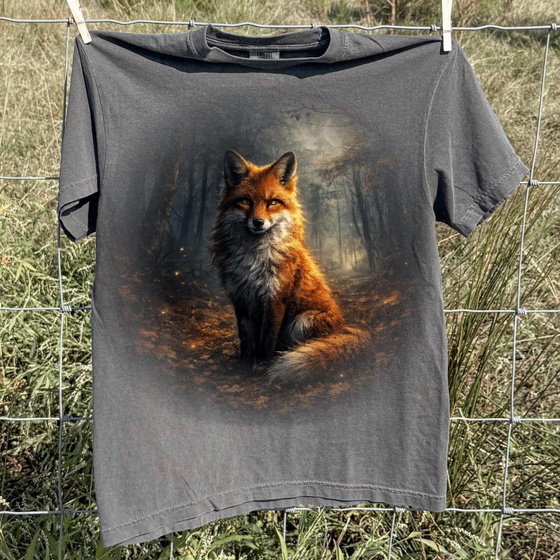 May include: A dark grey t-shirt displaying a detailed illustration of a fox with orange and white fur, set against a forest backdrop. The fox is sitting on a bed of leaves. The t-shirt is hanging on a clothesline.