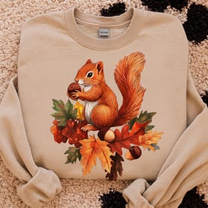 May include: A beige crewneck sweatshirt featuring a graphic illustration of a brown squirrel holding an acorn. The squirrel is surrounded by a cluster of colorful autumn leaves in shades of orange, yellow, and red, along with several scattered acorns.