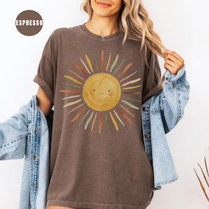 May include: An espresso-colored short-sleeve t-shirt featuring a central graphic of a smiling yellow sun with radiating brushstroke rays in shades of orange, yellow, and sage green. The text ESPRESSO appears in a brown circle in the upper left corner.