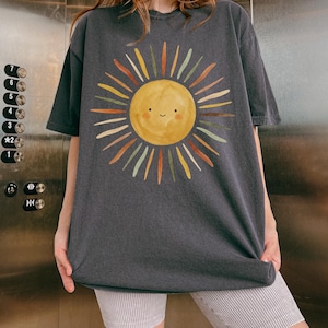 May include: A charcoal gray short-sleeve t-shirt featuring a large, centered graphic of a yellow sun with a simple smiling face. The sun has radiating rays in various muted tones including orange, green, brown, and cream, creating a watercolor effect.