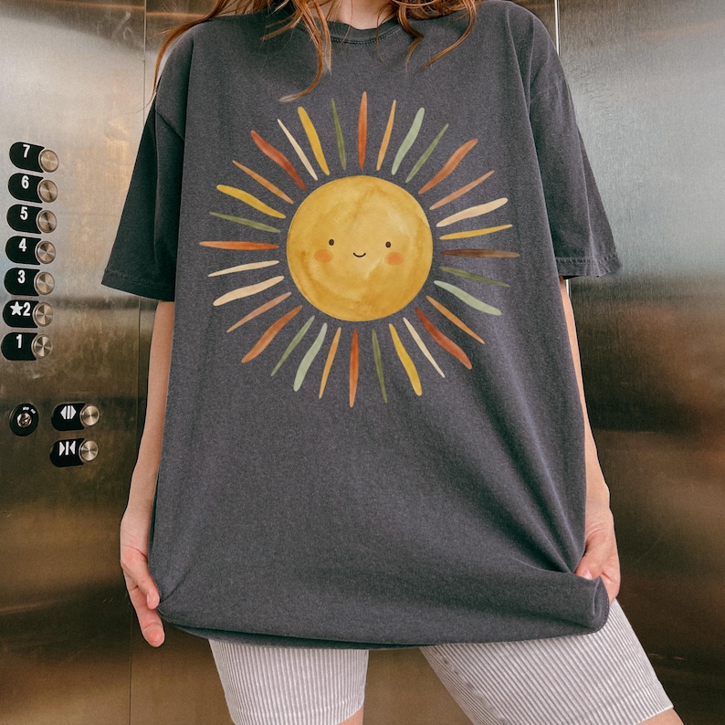 May include: A charcoal gray short-sleeve t-shirt featuring a large, centered graphic of a yellow sun with a simple smiling face. The sun has radiating rays in various muted tones including orange, green, brown, and cream, creating a watercolor effect.