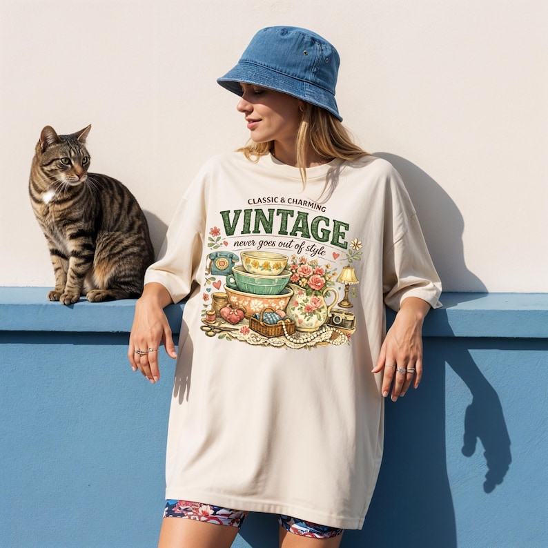 May include: A beige t-shirt featuring a graphic print of stacked bowls, a floral pitcher, a rotary phone, and a basket with the text "CLASSIC & CHARMING VINTAGE never goes out of style". The shirt is paired with a blue denim bucket hat.