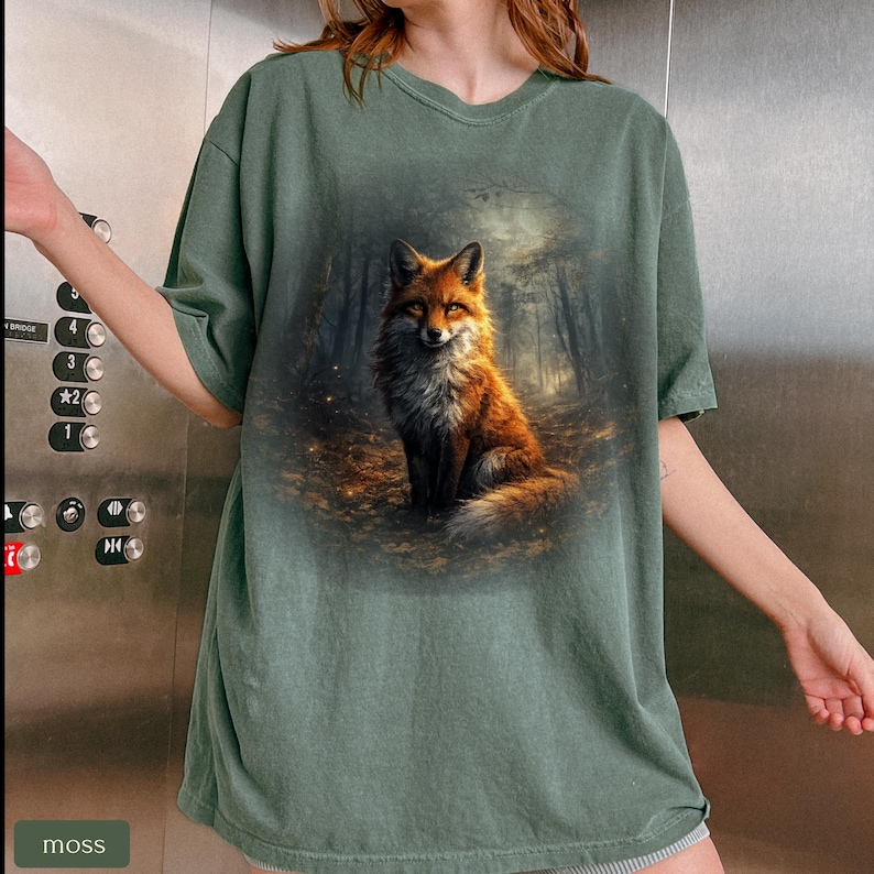 May include: A moss green t-shirt with a detailed illustration of a fox in a forest. The fox has orange and white fur, and the forest background is dark with tall trees. The word "moss" is in a small box in the lower left corner.