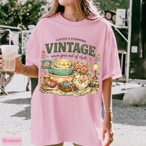 May include: A light pink t-shirt featuring a graphic print with the text "CLASSIC & CHARMING VINTAGE never goes out of style". The illustration includes stacked bowls, a floral pitcher, a rotary phone, a lamp, and sewing supplies.