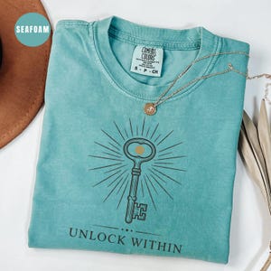 May include: Seafoam green Comfort Colors t-shirt with a key graphic and the words "UNLOCK WITHIN". The key design has a gold-colored center and radiating lines. A gold necklace with a pendant is draped across the shirt.
