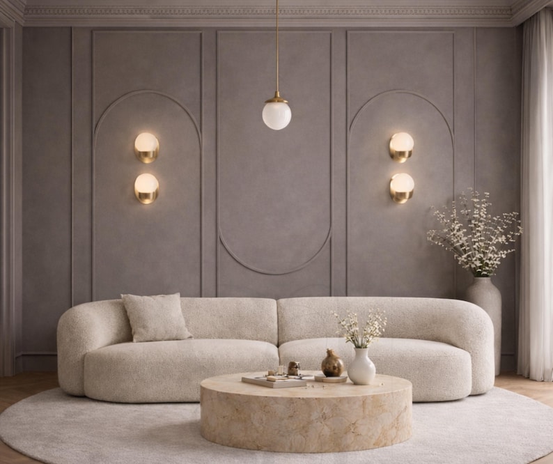 May include: A modern living room with a beige boucle sofa, a round stone coffee table, and a gray rug. The wall is a muted gray with decorative paneling and gold sconces. A pendant light hangs above the coffee table.