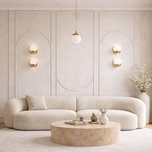 May include: A modern living room with a cream-colored curved sofa, a round coffee table, and a cream-colored rug. The walls are a light beige with decorative paneling and gold sconces. A white vase with flowers sits on the right.