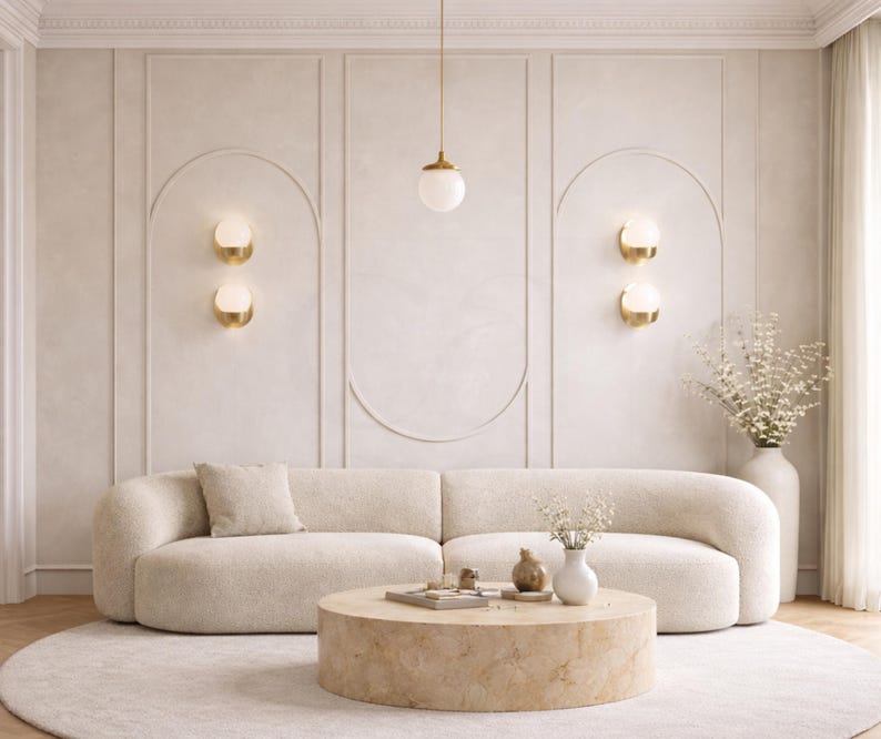 May include: A modern living room with a cream-colored curved sofa, a round coffee table, and a cream-colored rug. The walls are a light beige with decorative paneling and gold sconces. A white vase with flowers sits on the right.