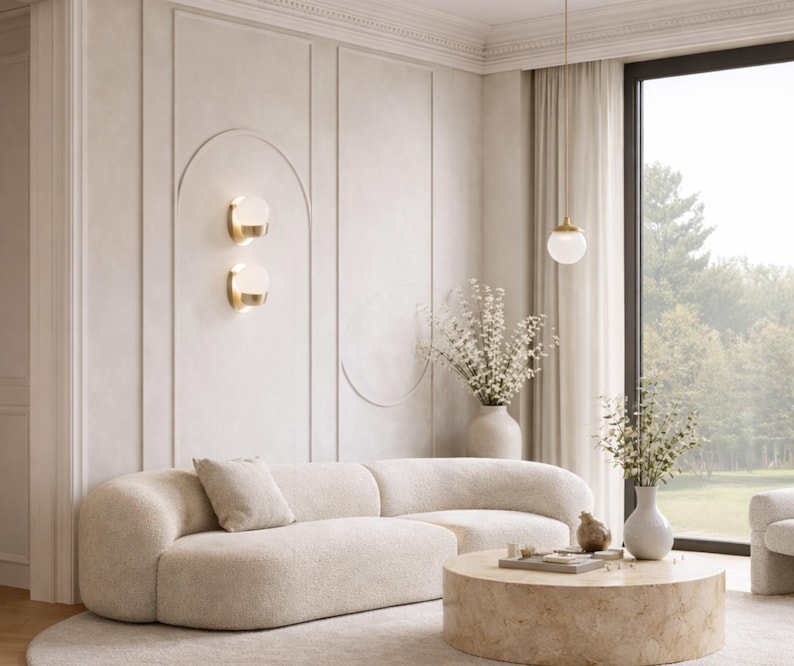 May include: A modern living room with a curved, cream-colored sofa, a round coffee table, and a large window. The walls are a light neutral color with decorative paneling and two wall sconces. A vase of flowers sits on the coffee table.