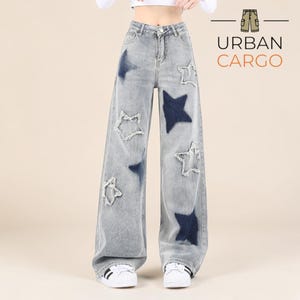 May include: Light wash denim jeans with a wide leg cut. The jeans have dark blue star patches and star-shaped cutouts. The text "URBAN CARGO" is displayed in a logo on the upper right corner.