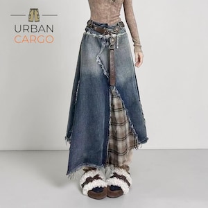 Peut inclure: Longue jupe en jean délavé avec un design asymétrique et vieilli. La jupe présente un panneau en tartan marron et une ceinture en cuir marron avec des détails métalliques décoratifs. Le logo "URBAN CARGO" est visible.