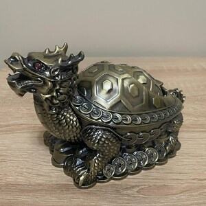 May include: A bronze-toned dragon-headed turtle figurine featuring a detailed shell with hexagonal patterns, scales on the neck and legs, and a base adorned with a row of circular coins. The creature has small red eyes and an open mouth.