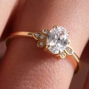 Pode incluir: Um anel de ouro com uma pedra preciosa oval e transparente. O anel tem um design delicado com pequenas pedras redondas e uma cravação floral. A banda é de ouro fino e polido.