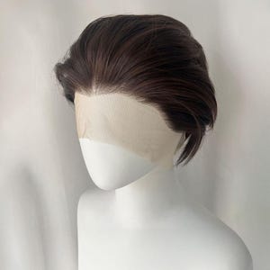 Op de afbeelding: Een donkerbruine pruik met een kanten voorkant wordt getoond op een witte mannequinhoofd. De pruik heeft een korte, gelaagde stijl met een zijscheiding. Het haar is gestyled met een lichte golf en is netjes gekamd.