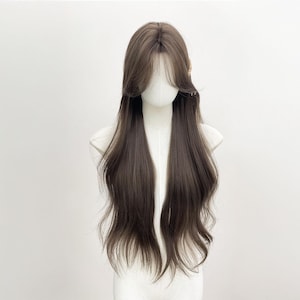 Op de afbeelding: Een lange, bruine pruik met een natuurlijk ogende haarlijn en zijwaarts geveegde pony. De pruik is gestyled met lange, vloeiende lagen en een lichte golf aan de uiteinden. Een klein, goudkleurig haaraccessoire is zichtbaar bij de kruin. De pruik wordt getoond op een mannequinhoofd.