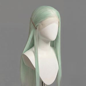 Puede incluir: Una peluca verde menta con cabello largo y liso se muestra sobre una cabeza y hombros de maniquí blancos. Una horquilla dorada asegura el cabello en un lado. El frente de encaje de la peluca es visible, y el fondo es gris liso.