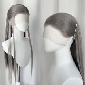 Pode incluir: Uma peruca longa cinza prateada com uma risca lateral e madeixas longas e lisas. É exibida em uma cabeça de manequim branca, mostrando a frente de renda e a linha do cabelo natural. Uma presilha prateada prende o cabelo de um lado.