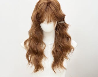 May include: A light brown wig with long, wavy hair and bangs, displayed on a mannequin head. A small silver hair clip secures a section of the hair. The wig has a natural look and is styled with soft waves.