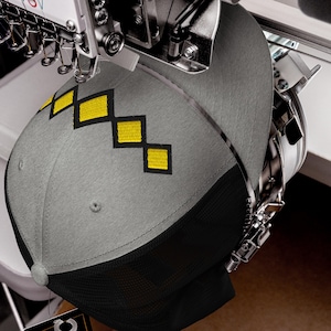 Op de afbeelding: Een grijze baseballcap met een zwarte mesh achterkant wordt geborduurd door een machine. De cap heeft een patroon van gele ruiten met zwarte omlijning. De borduurmachine is zilverkleurig en heeft meerdere naalden.