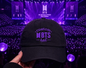 Può includere: Un cappellino da baseball nero con ricamo viola sul davanti. Il testo recita custom BTS hat, accompagnato dal logo BTS. Il design include piccoli accenti a forma di stella sopra il testo e linee orizzontali ai lati della parola hat.