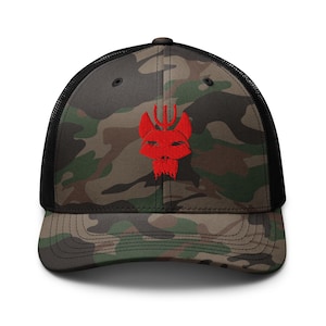 May include: Camouflage trucker hat with a black mesh back and a curved brim. The front panel features a red embroidered design of a stylized wolf head with a trident. The hat is a mix of green, brown, and black.